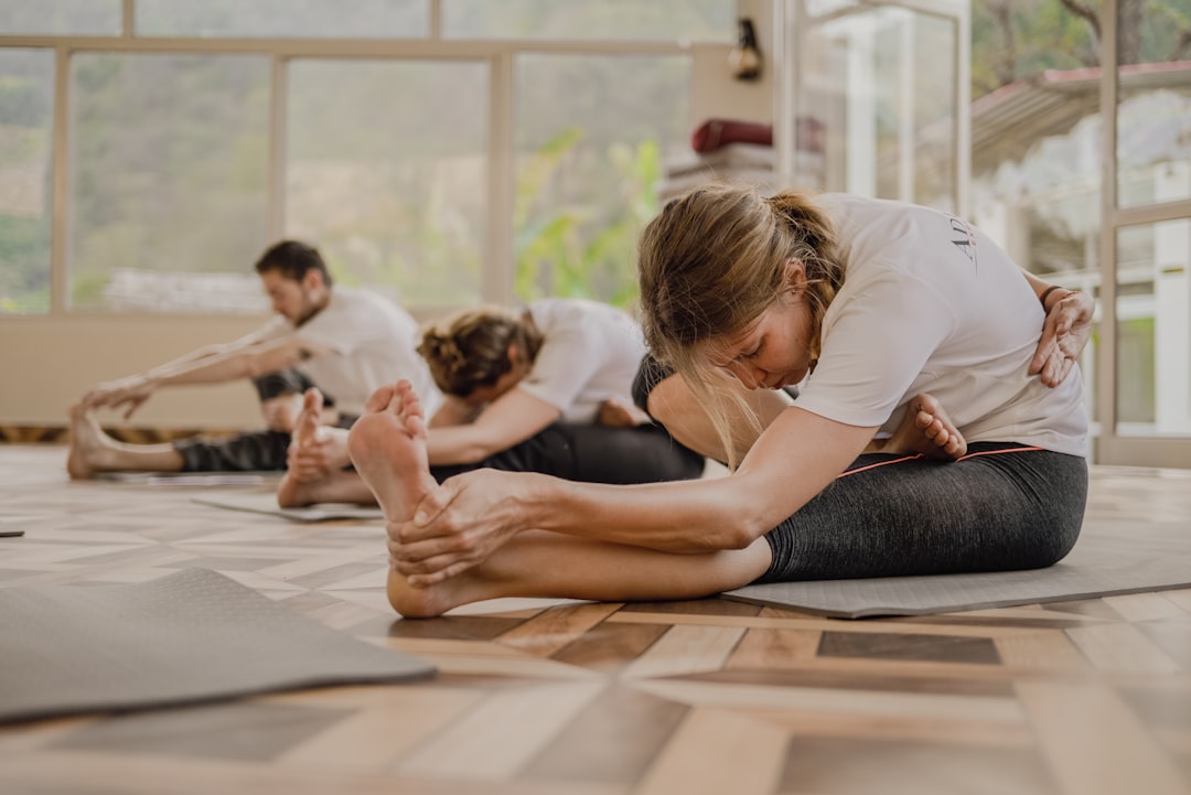 Drei Personen sitzen auf Yogamatten in einem Raum und strecken sich nach vorne, um ihre Zehen in einer Gruppenübungsstunde mit Schwerpunkt auf Pilates-Übungen und Stärkung des Kerns zu berühren.